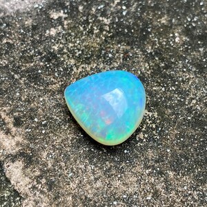 May include: A teardrop-shaped opal gemstone with a light blue and green iridescent sheen. The opal is set against a rough, gray background.