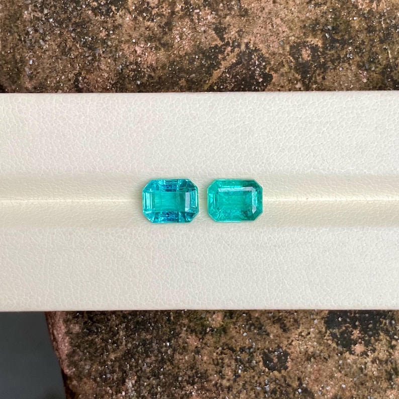 May include: Two rectangular, faceted gemstones with a vibrant teal color. The gems are displayed on a white surface, showcasing their clarity and brilliance. The gemstones are likely for jewelry making or collecting.