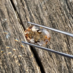 May include: A light brown oval-shaped gemstone with facets, held by tweezers against a wooden background.