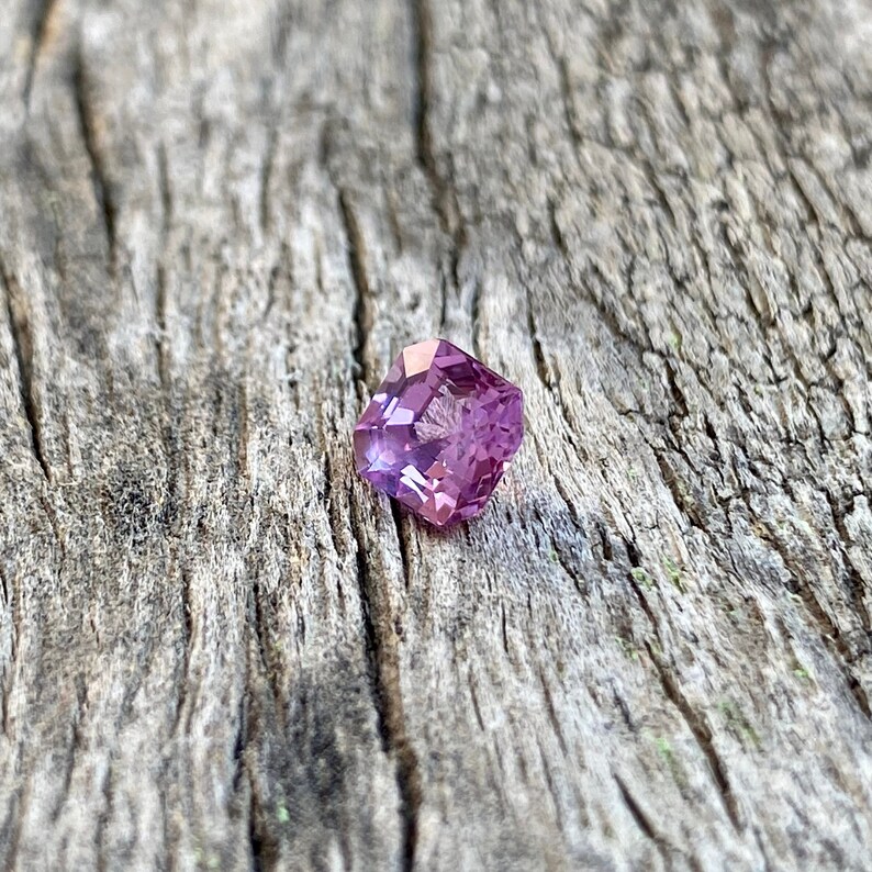 Puede incluir: Una piedra preciosa facetada de forma cuadrada con un tono p&uacute;rpura. La piedra preciosa se coloca sobre un fondo de madera r&uacute;stico.