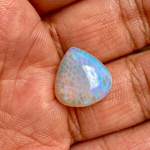 May include: A teardrop-shaped opal gemstone with a white base and iridescent blue and purple hues. The opal has a textured surface with a pattern resembling scales.