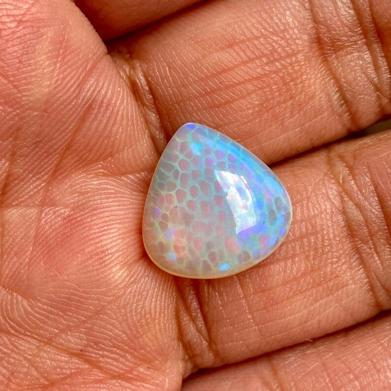 May include: A teardrop-shaped opal gemstone with a white base and iridescent blue and purple hues. The opal has a textured surface with a pattern resembling scales.