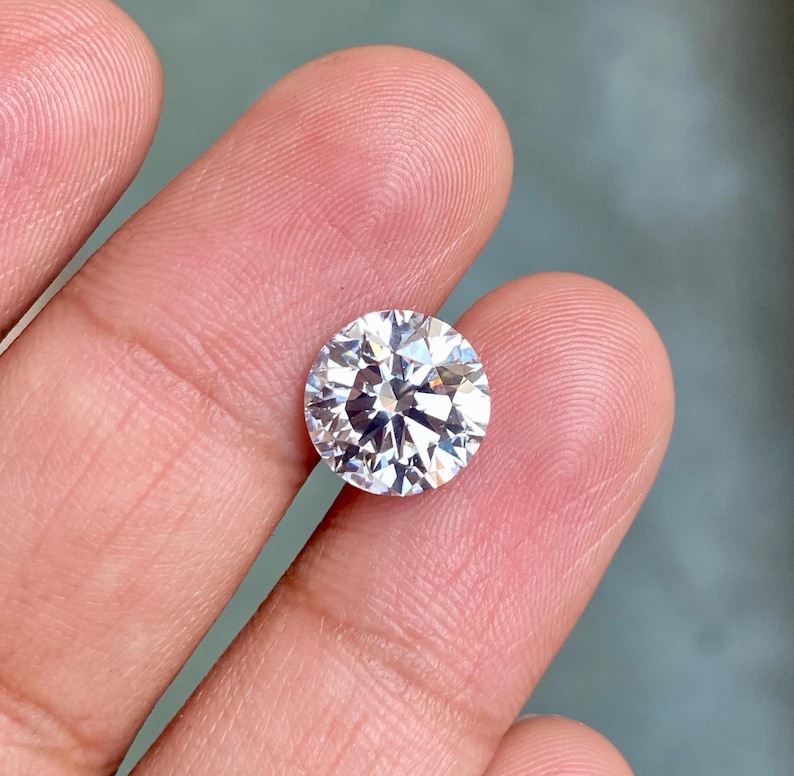 May include: A large, clear, round, faceted gemstone with a brilliant cut. The gemstone is held in a person's hand.