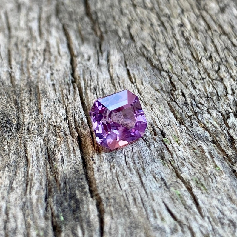 Puede incluir: Una piedra preciosa cuadrada de color p&uacute;rpura con un corte facetado. La piedra preciosa est&aacute; colocada sobre un fondo de madera r&uacute;stico.