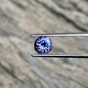 May include: A round, faceted, purple gemstone with a blue hue is held in tweezers. The gemstone is a vibrant colour and has a high level of brilliance.