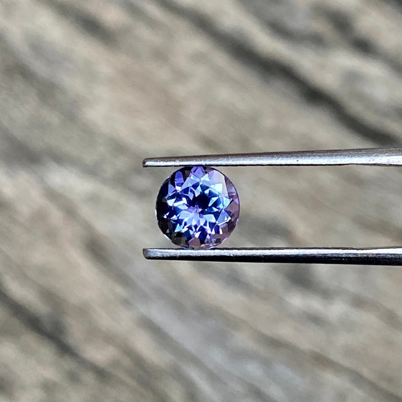 May include: A round, faceted, purple gemstone with a blue hue is held in tweezers. The gemstone is a vibrant colour and has a high level of brilliance.