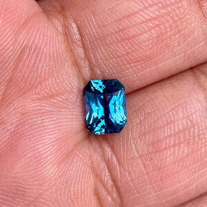 May include: A vibrant blue gemstone with a rectangular emerald cut. The gemstone is set in a bezel setting and has a high polish.