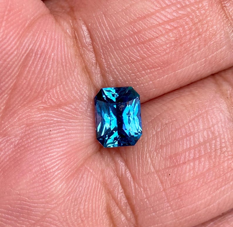 May include: A vibrant blue gemstone with a rectangular emerald cut. The gemstone is set in a bezel setting and has a high polish.