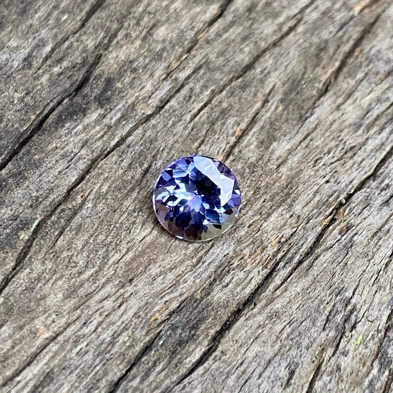 May include: A round, faceted, purple gemstone with a blue hue. The gemstone is set on a wooden surface.