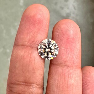May include: A clear round diamond gemstone with facets, held between two fingers.