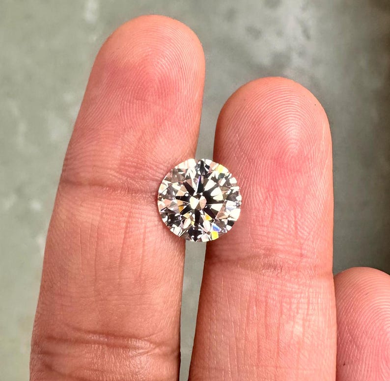 May include: A clear round diamond gemstone with facets, held between two fingers.