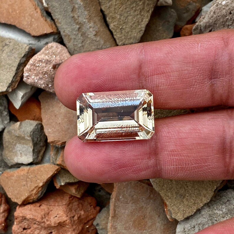 May include: A light brown, rectangular, faceted gemstone with a slightly darker brown center. The gemstone is held between two fingers.