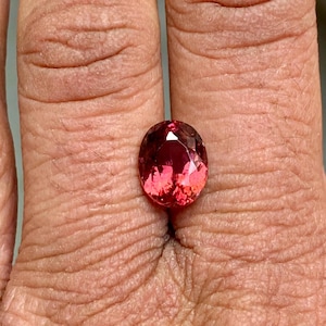 K&ouml;nnte beinhalten: Ovaler, facettierter Edelstein in rosafarbenem und rotem Farbton. Der Edelstein ist in einem Ring gefasst und wird an einem Finger getragen.