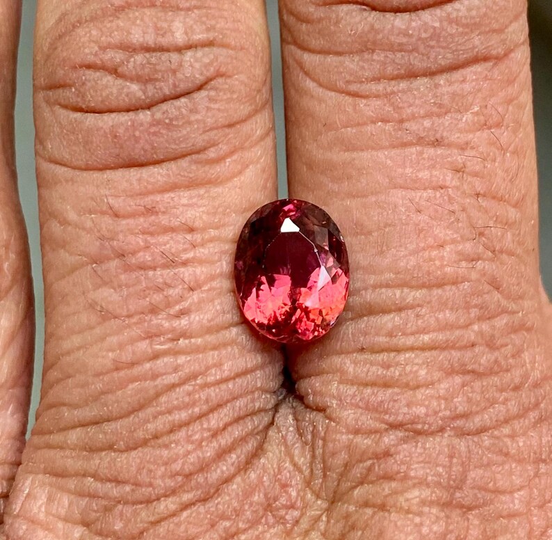 K&ouml;nnte beinhalten: Ovaler, facettierter Edelstein in rosafarbenem und rotem Farbton. Der Edelstein ist in einem Ring gefasst und wird an einem Finger getragen.