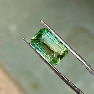 May include: A green gemstone with a rectangular emerald cut, held in tweezers. The gemstone has a slight color gradient from a darker green to a lighter green.
