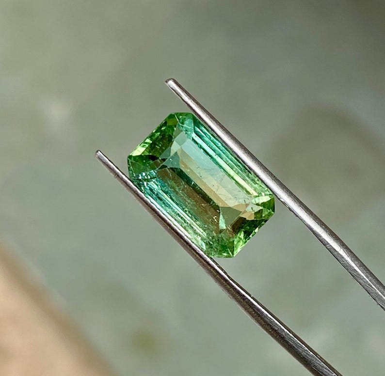 May include: A green gemstone with a rectangular emerald cut, held in tweezers. The gemstone has a slight color gradient from a darker green to a lighter green.