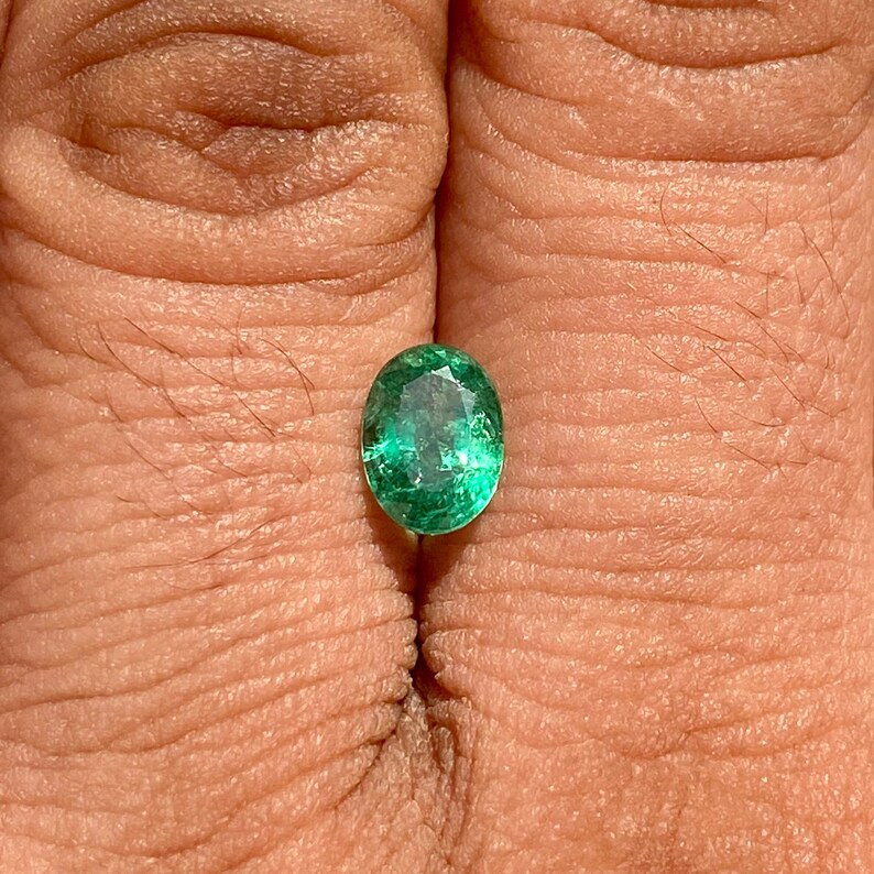 May include: A vibrant green oval emerald gemstone, set against a neutral background.