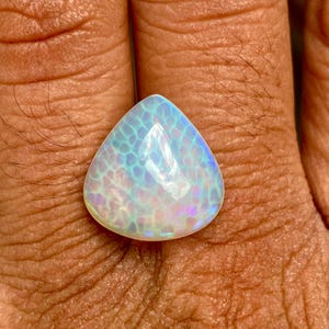 May include: A teardrop-shaped opal gemstone with a white base and iridescent blue and purple hues. The opal has a unique pattern of small, white, and blue spots.