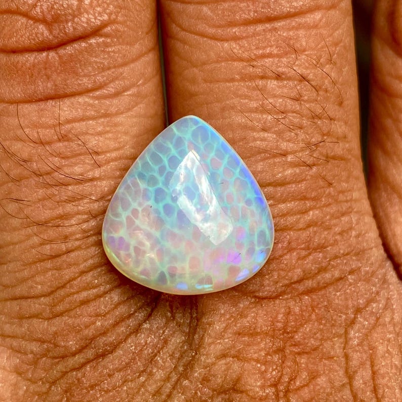 May include: A teardrop-shaped opal gemstone with a white base and iridescent blue and purple hues. The opal has a unique pattern of small, white, and blue spots.