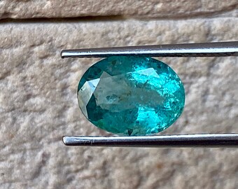 May include: A vibrant green oval-shaped gemstone, likely an emerald, held in tweezers against a stone background.