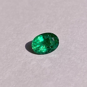 May include: A vibrant green emerald gemstone, cut in an oval shape, is displayed on a white surface.