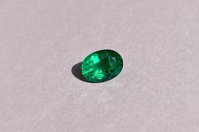 May include: A vibrant green emerald gemstone, cut in an oval shape, is displayed on a white surface.