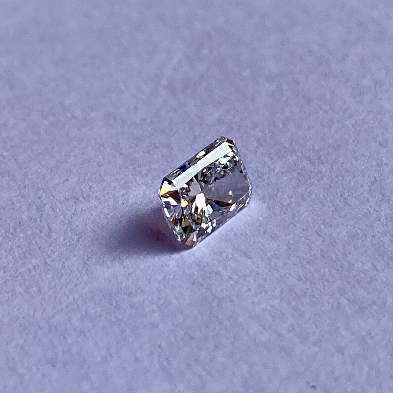以下が含まれることがあります： 明るいカットが施された、透明な長方形のファセットカットの宝石。宝石は薄い紫色の背景に置かれています。