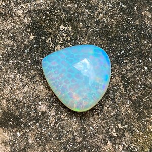 May include: A teardrop-shaped opal gemstone with a light blue and iridescent color. The opal has a textured surface and is set against a rough, gray background.