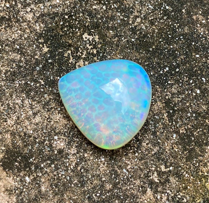 May include: A teardrop-shaped opal gemstone with a light blue and iridescent color. The opal has a textured surface and is set against a rough, gray background.