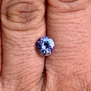 May include: A round, faceted gemstone with a purple and blue colour gradient. The gemstone is set in a bezel setting and is being held between two fingers.