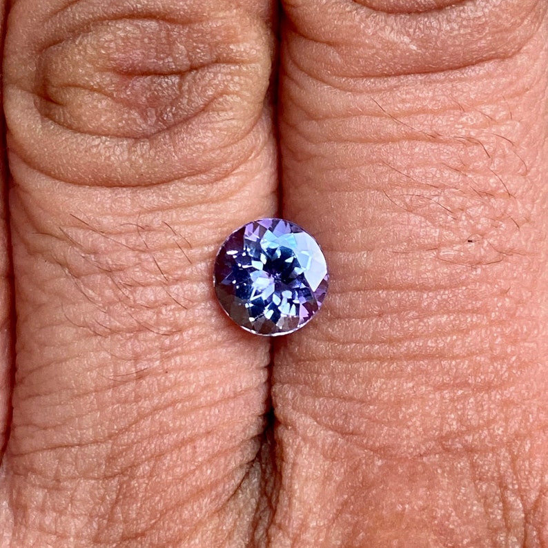 May include: A round, faceted gemstone with a purple and blue colour gradient. The gemstone is set in a bezel setting and is being held between two fingers.