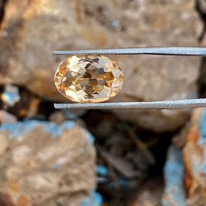 May include: A light brown oval-shaped gemstone with facets, held by tweezers against a rocky background.