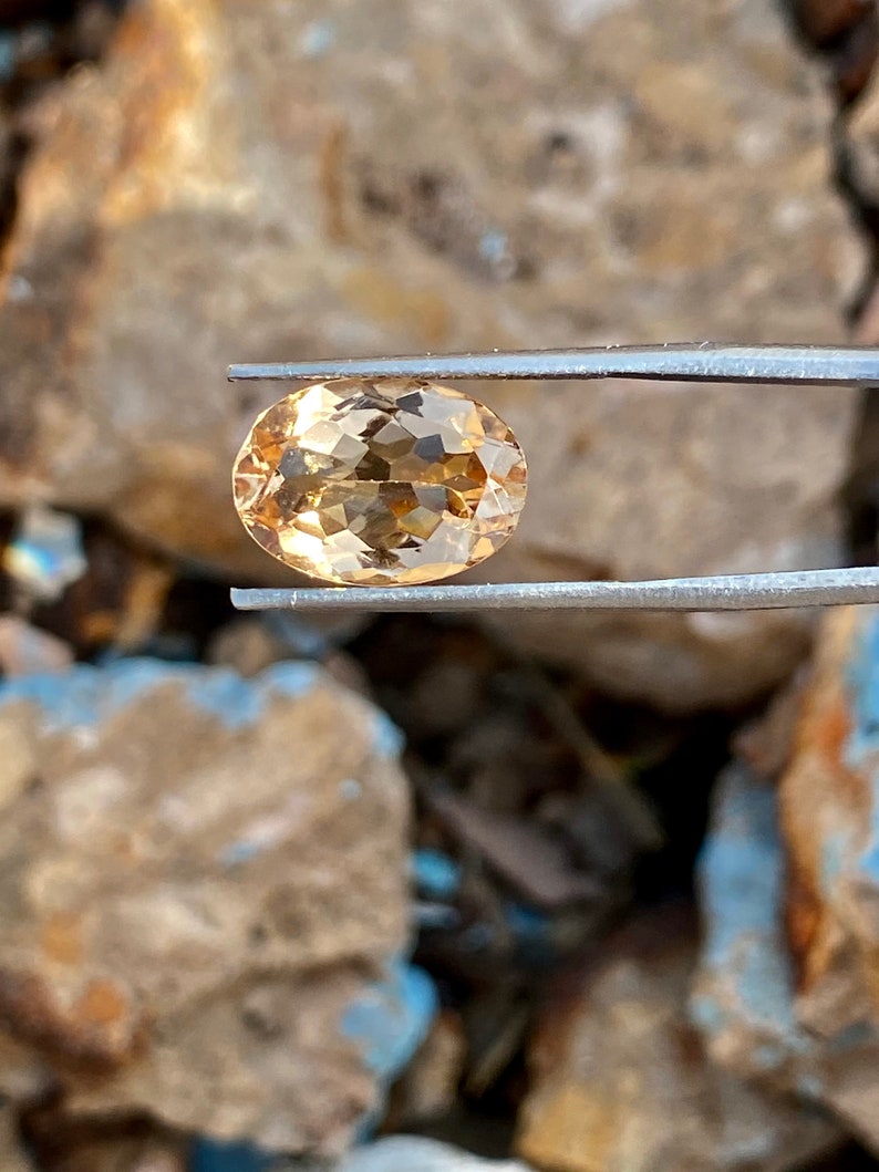 May include: A light brown oval-shaped gemstone with facets, held by tweezers against a rocky background.