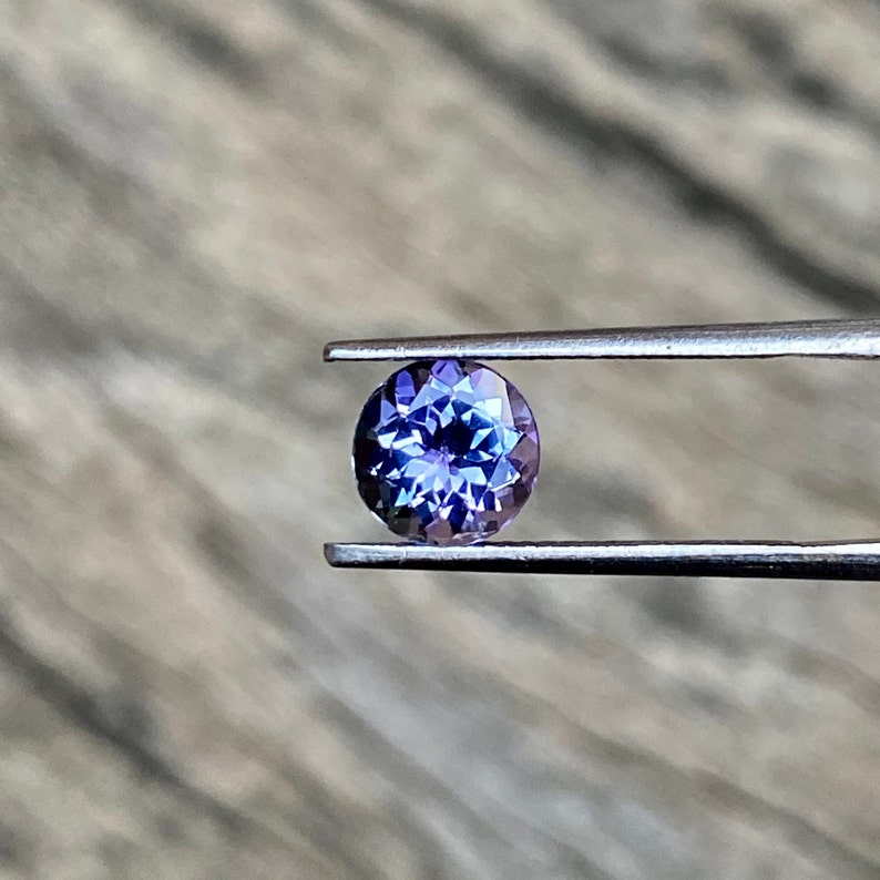 May include: A round, purple gemstone with a faceted cut, held by tweezers against a blurred wooden background.