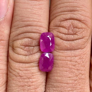 May include: Two cushion-cut ruby gemstones with a deep pink-red hue. The faceted stones are displayed between fingers, highlighting their polished surfaces and internal sparkle. The rubies are ready to be set into jewelry, showcasing their vibrant colour.