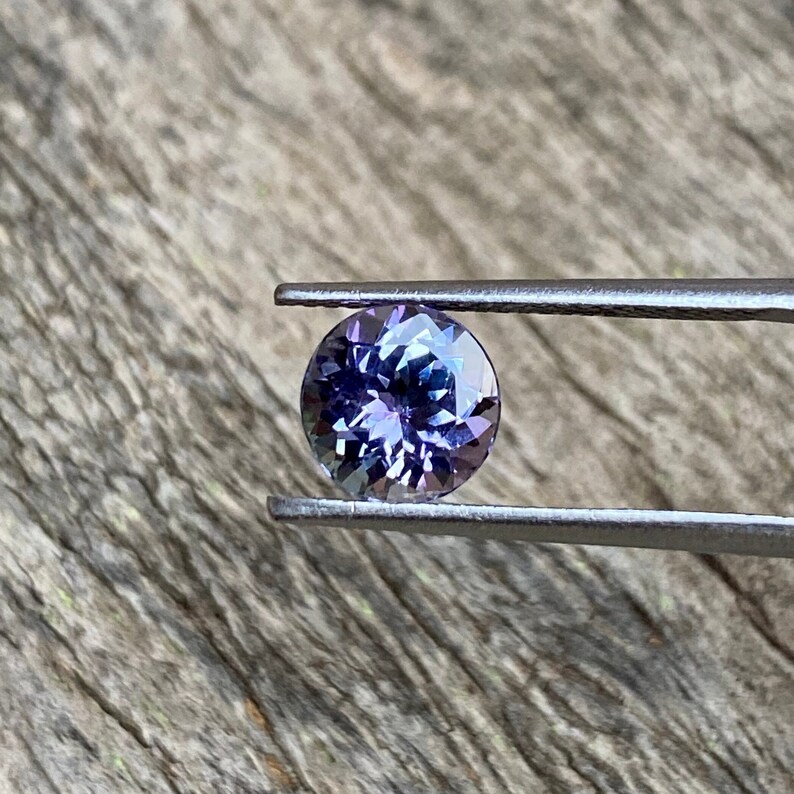 May include: A round, faceted gemstone with a purple and blue hue. The gemstone is held in place by a pair of tweezers.