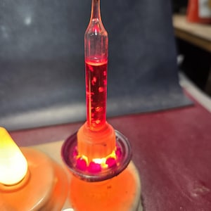 May include: A cylindrical glass bubble light featuring a vibrant red liquid interior with suspended particles. The light is mounted on a circular base with a glowing orange light source at the bottom, creating a bright illumination effect.