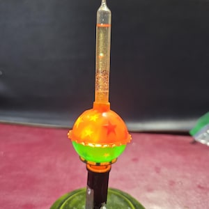 May include: A decorative bubble light featuring a clear glass tube filled with orange liquid and glitter, topped with a spherical orange and green plastic base decorated with red and yellow star patterns, mounted on a dark green circular stand.
