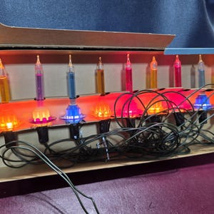 May include: A string of nine multi-colored bubble lights with a dark green cord, housed in a cardboard box. The glass bulbs feature liquid-filled tubes in yellow, purple, blue, orange, pink, and red, each set in a plastic base.