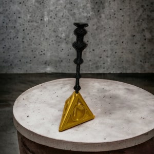 May include: A black and gold decorative object. The object has a gold triangular base with a symbol and a black, ornate vertical element. The object is displayed on a round, light-colored surface against a gray background.