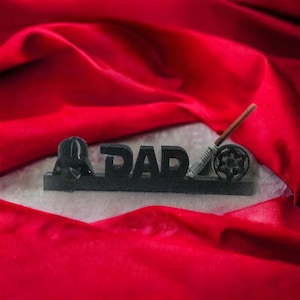 May include: A black decorative piece with the word "DAD" in block letters, featuring a Darth Vader helmet, a lightsaber, and a Galactic Empire symbol. The piece is set against a red fabric background.