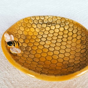 Op de afbeelding: Een klein keramisch schaaltje in de vorm van een honingraat met een bijenfiguur aan de zijkant. Het schaaltje is geel met zwarte lijnen en is perfect voor het bewaren van kleine voorwerpen.