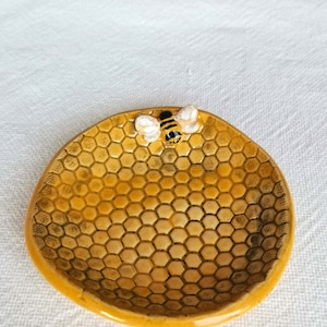 Op de afbeelding: Een klein, geel keramisch schaaltje in de vorm van een honingraat met een bij-figuur erop. Het schaaltje is perfect om kleine spulletjes zoals sieraden of sleutels in te bewaren.