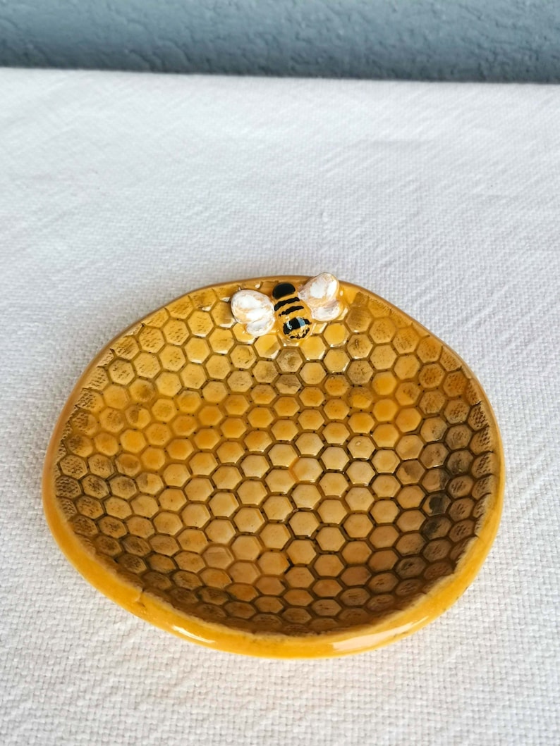 Op de afbeelding: Een klein, geel keramisch schaaltje in de vorm van een honingraat met een bij-figuur erop. Het schaaltje is perfect om kleine spulletjes zoals sieraden of sleutels in te bewaren.