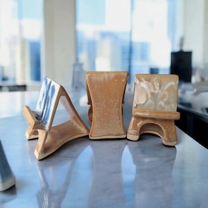 Puede incluir: Tres soportes de teléfono de cerámica en fila. Los soportes son en tonos beige y marrón, con un diseño azul y blanco en uno. Los soportes tienen diferentes formas, incluyendo un marco en A y un diseño rectangular.