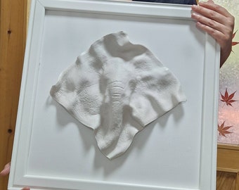 May include: A white plaster cast of a baby's handprint in a white frame. The handprint is in the shape of an elephant.