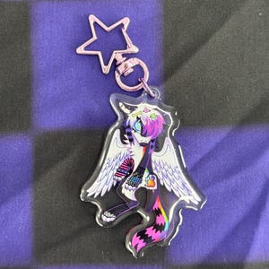 May include: A clear acrylic keychain featuring a stylized winged creature with a black and white body, purple and pink hair, and a rainbow tail. The keychain has a star-shaped clasp and a silver ring.