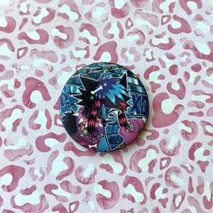 May include: A round button pin featuring a stylized cartoon character with black and blue fur, set against a brick wall background. The pin has a chain border and the letters "XD". The background is a pink and white leopard print.