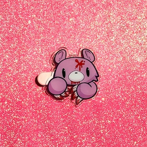 May include: A pink bear-shaped acrylic pin with a distressed design. The bear has a red star-shaped mark on its forehead and appears to be holding a torn, bloody object. The background is a sparkly pink surface.