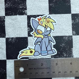 May include: A sticker of a cartoon pony with a light blue body, yellow hair, and large orange eyes. The pony has a cupcake and other food items on its head. The text "DERP XO" is on the pony's belly. The sticker is approximately 5 cm wide.